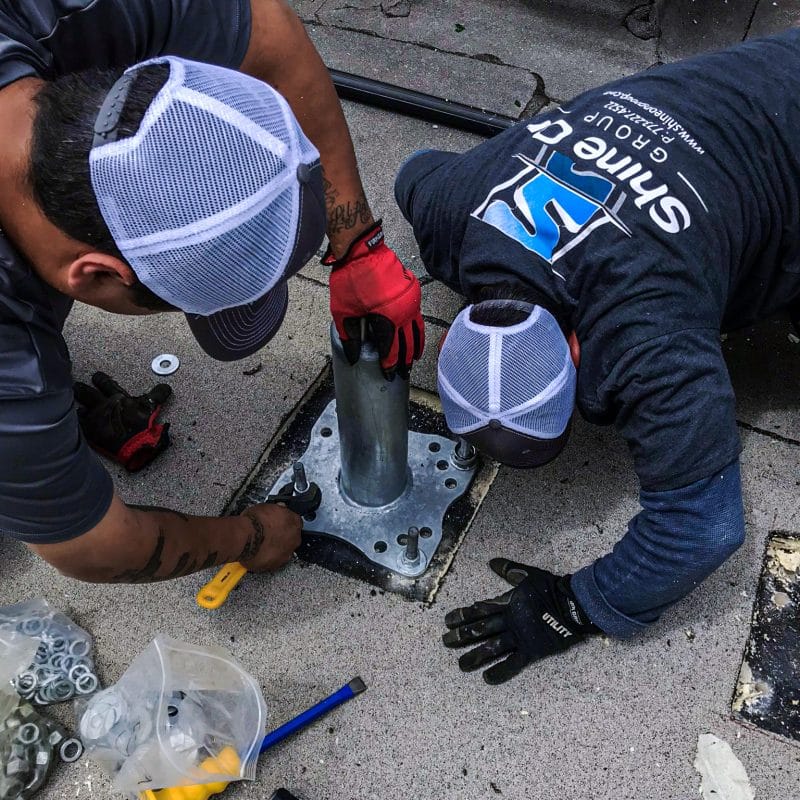 Two workers in "Shine Group" shirts install a metal pole base on a gray roof, using tools.